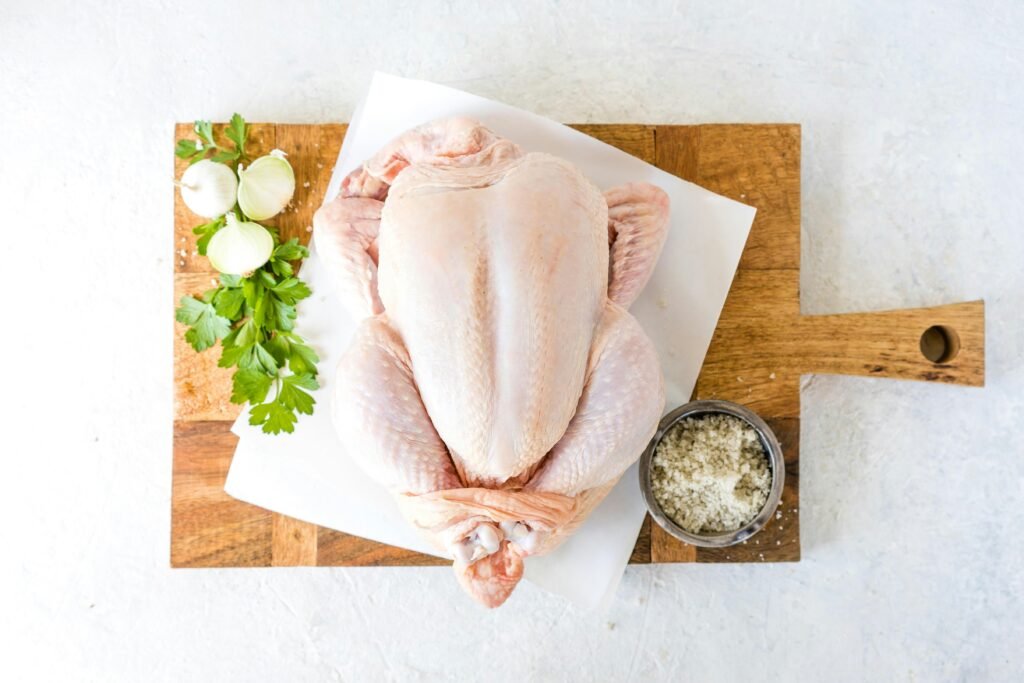 frango cru inteiro, em cima de uma tabua de corte , com algumas ervas aromaticas a decorar.
