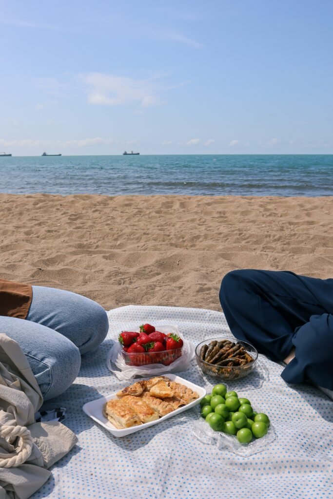 Duas pessoas a comer na praia, tem uma toalha estendida na areia com fruta e sandes, reforçando a ideia de levar comida para a praia em segurança