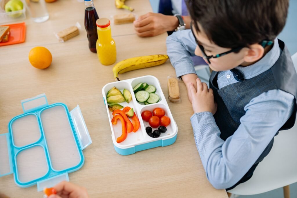Menino sentado a mesa com lancheira escolar a frente, com fruta e legumes , lanche saudável